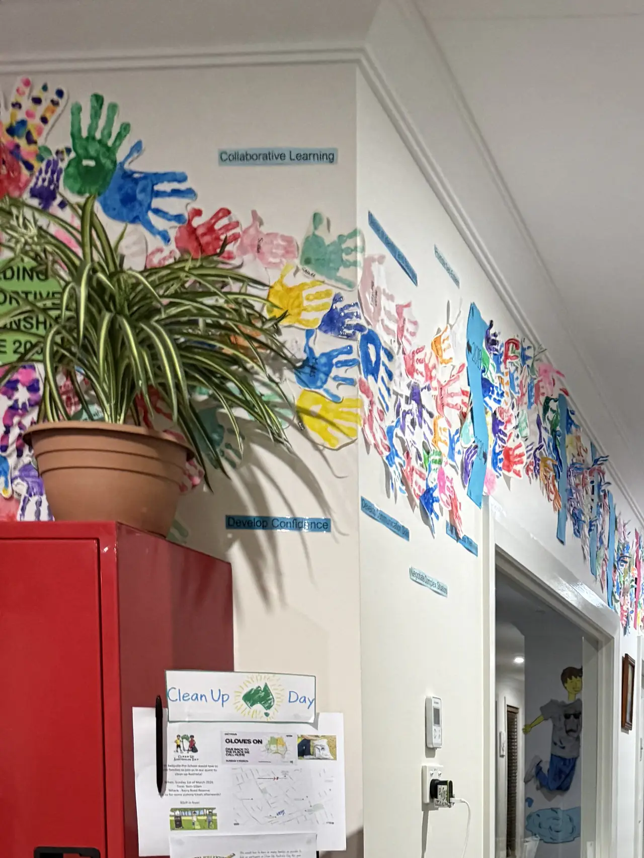 A corridor wall of children's handprint artwork above a red storage cabinet