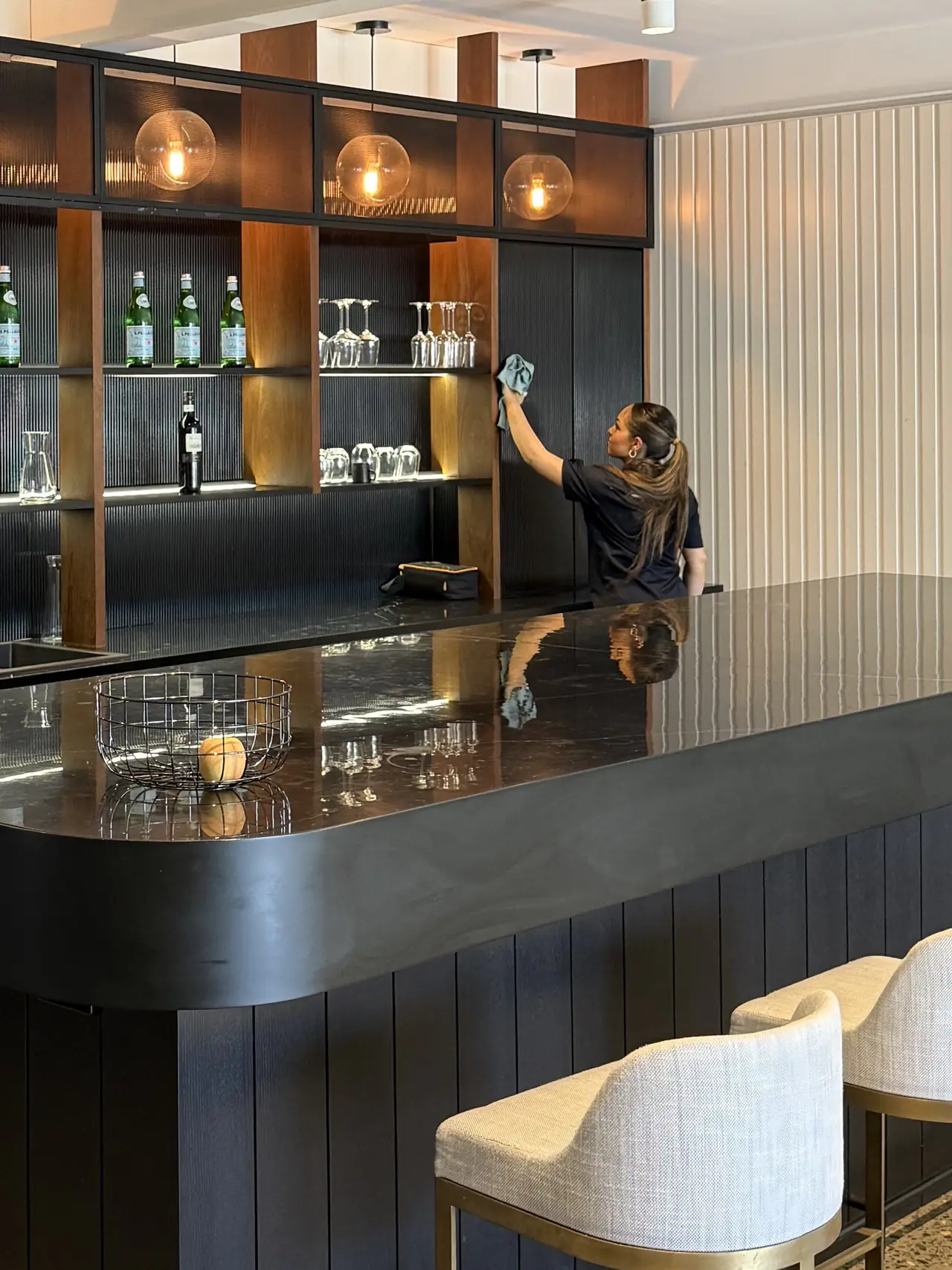 A cleaner polishing glassware on the back bar shelves above a wiped bar top and stools
